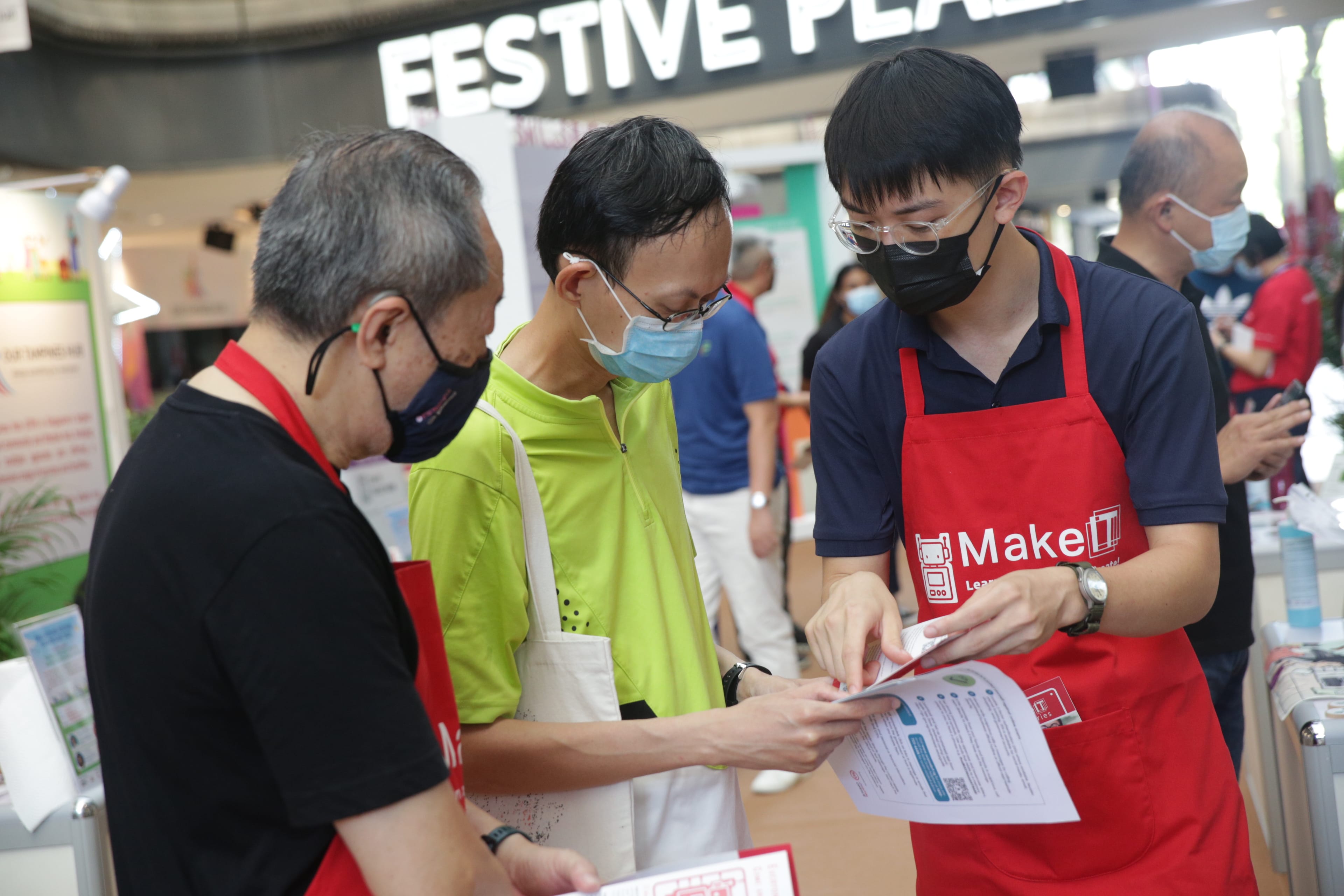 Individuals in an indoor setting are examining printed materials, with one wearing a "MakeIT" red apron.